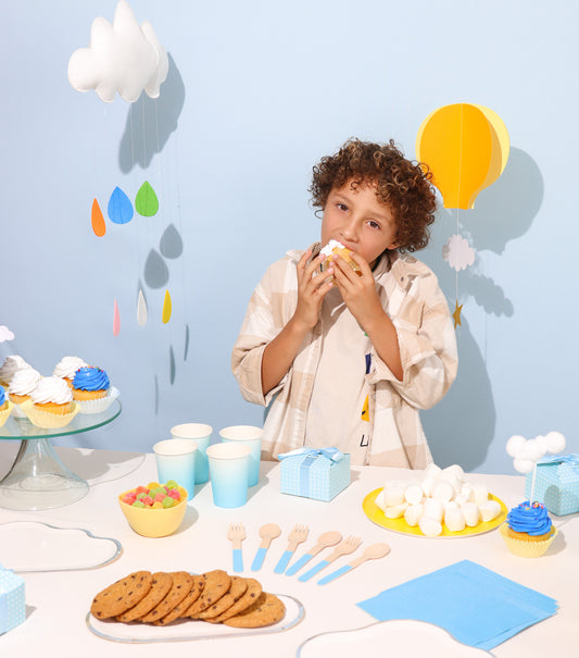 Child enjoying a cloud-themed party with cloud-shaped plates, yellow plates, cupcakes, and cookies. The scene is adorned with cloth cloud decorations and whimsical hot air balloons, creating a festive atmosphere of joy and celebration.